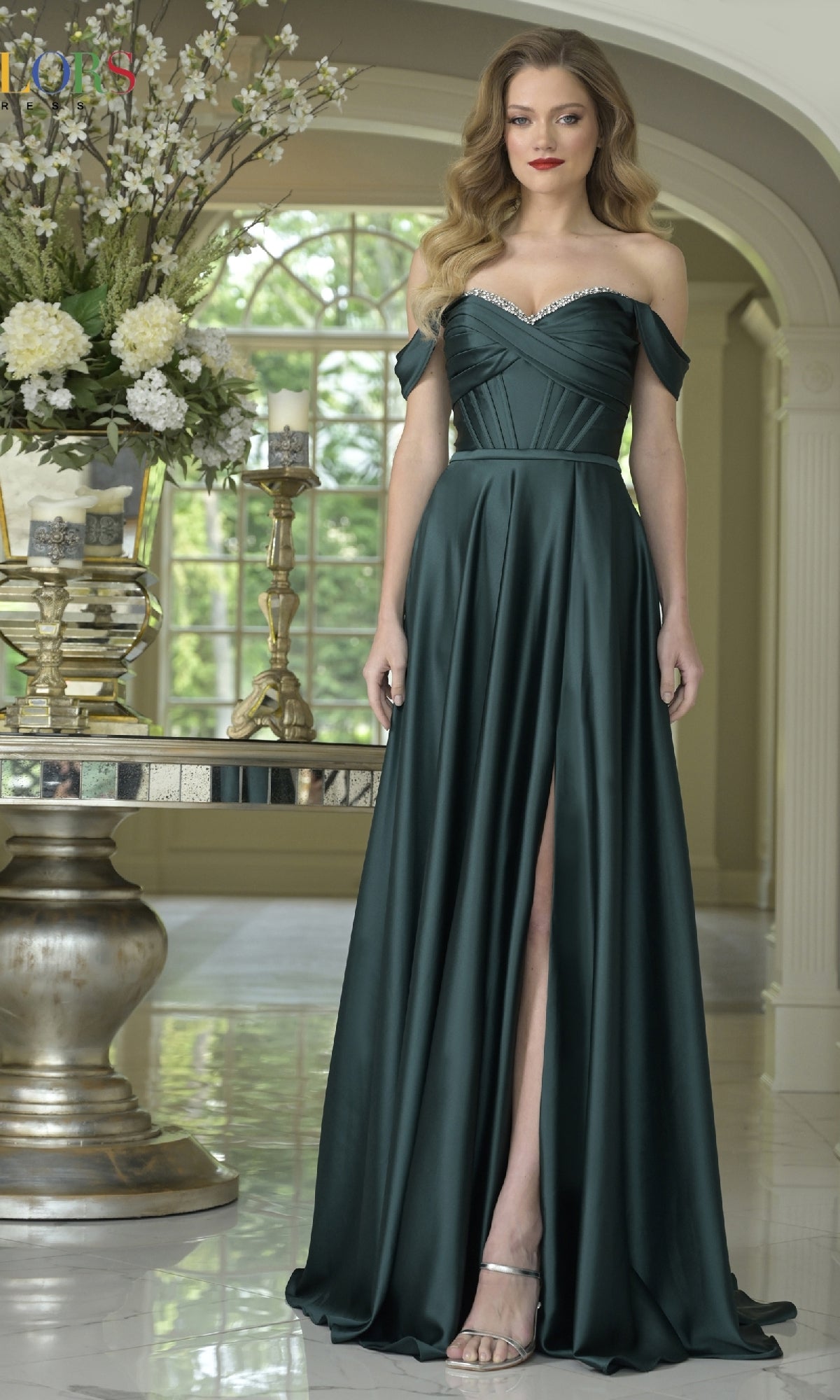 A woman in the Long Sweetheart Prom Dress: Colors Dress 3606, featuring a dark green off-the-shoulder design with high slit, stands indoors beside a table adorned with a large floral arrangement.